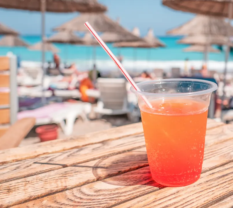 A plastic cup filled with an orange fizzy drink and a straw sits on a wooden table at a sunny beach bar, with blurred sun loungers, people, and straw umbrellas in the background.