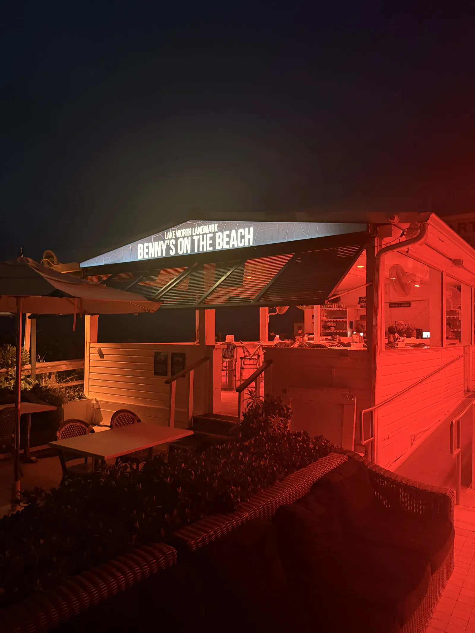 The neon Benny's On The Beach sign at night, red light across the empty tables below