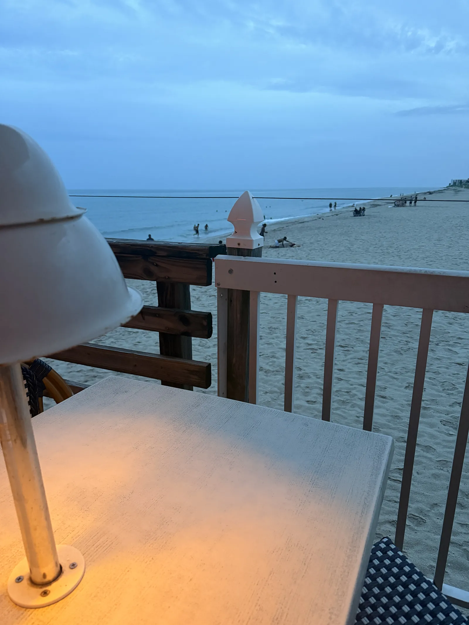 A lit table lamp at Benny's at dusk, the calm ocean and walkers on the sand beyond