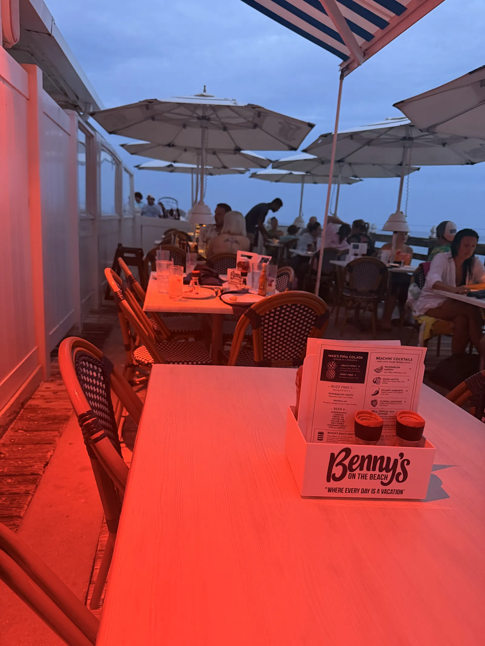 A Benny's on the Beach menu holder on the nearest table at dusk, diners under the umbrellas