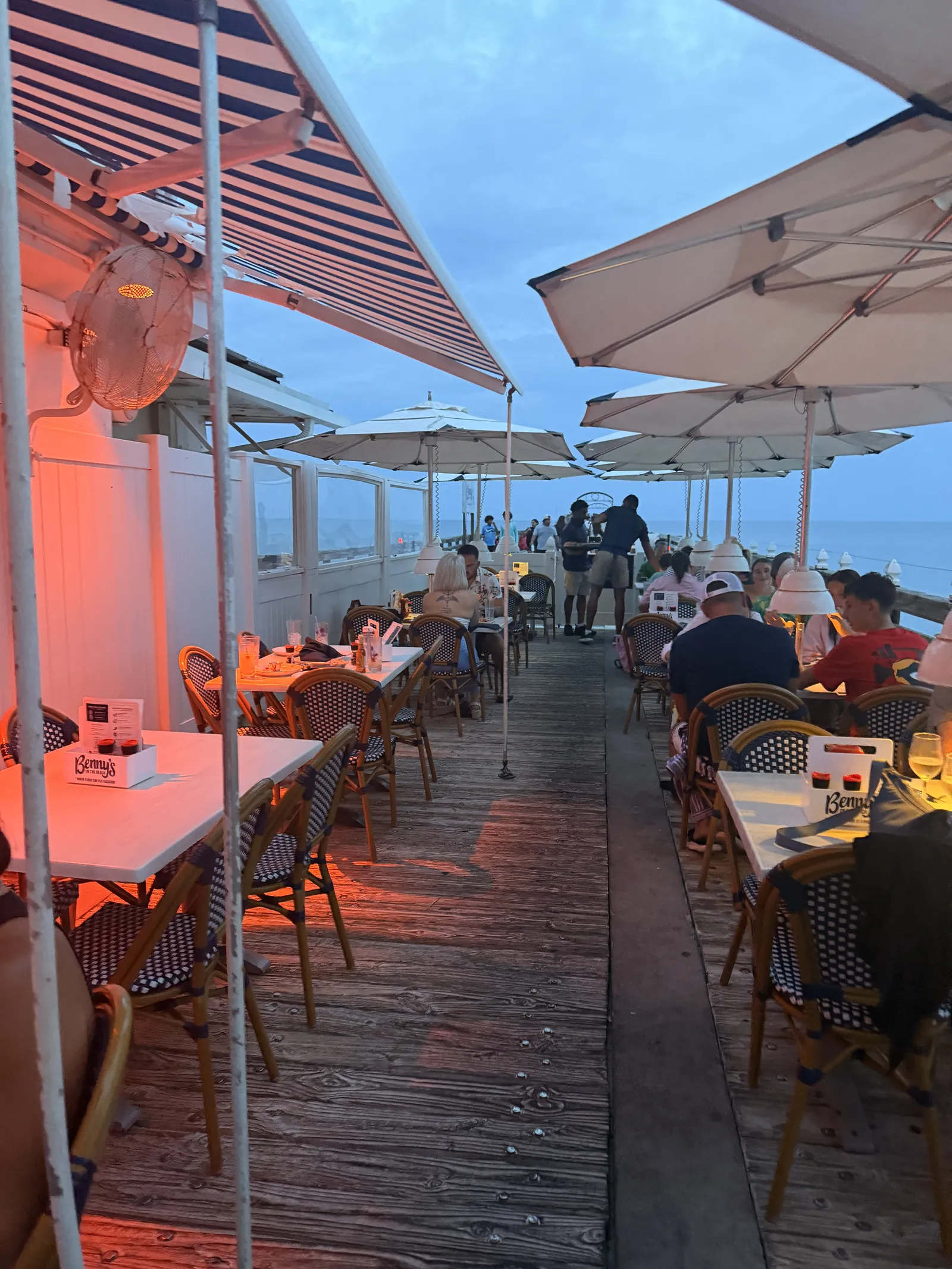 Diners under umbrellas on the deck at Benny's at dusk, warm lights and the ocean behind
