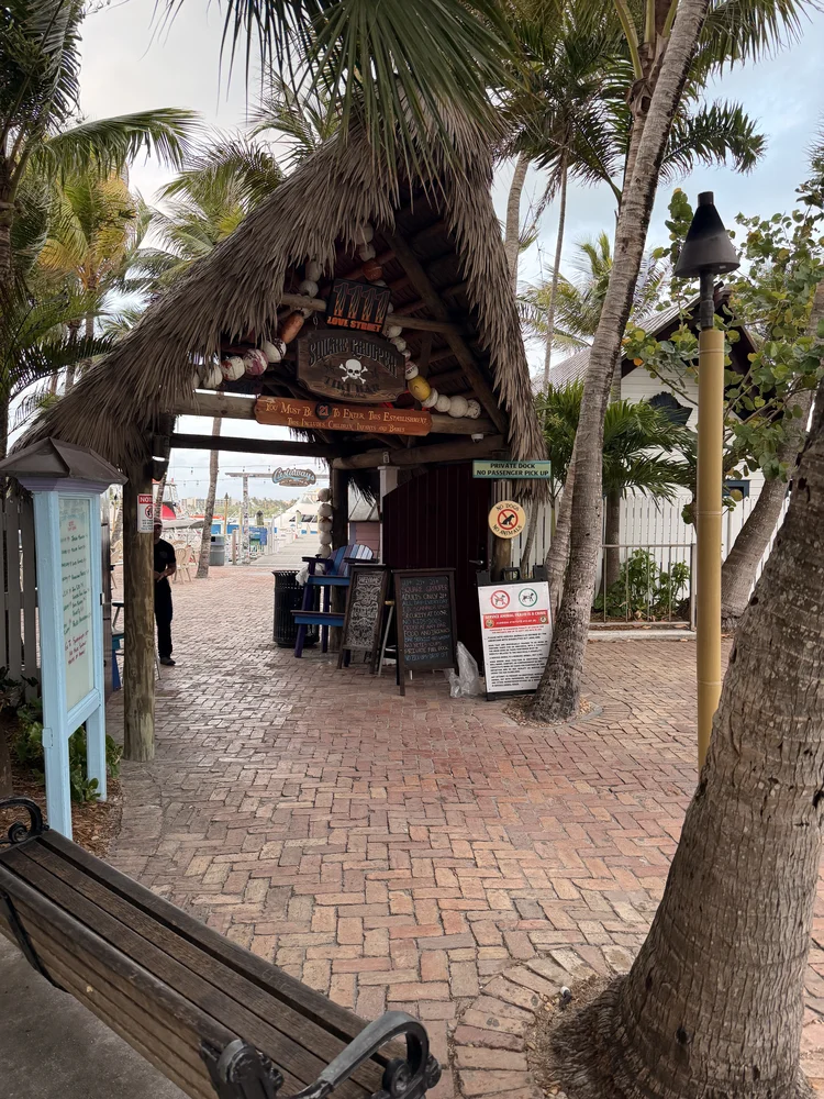 A thatched-roof entrance to a beachside restaurant, resembling the relaxed vibe of beach bars, features hanging decorations, chalkboard menus, and palm trees. A brick pathway leads toward the ocean, with a bench and lamp post in front.