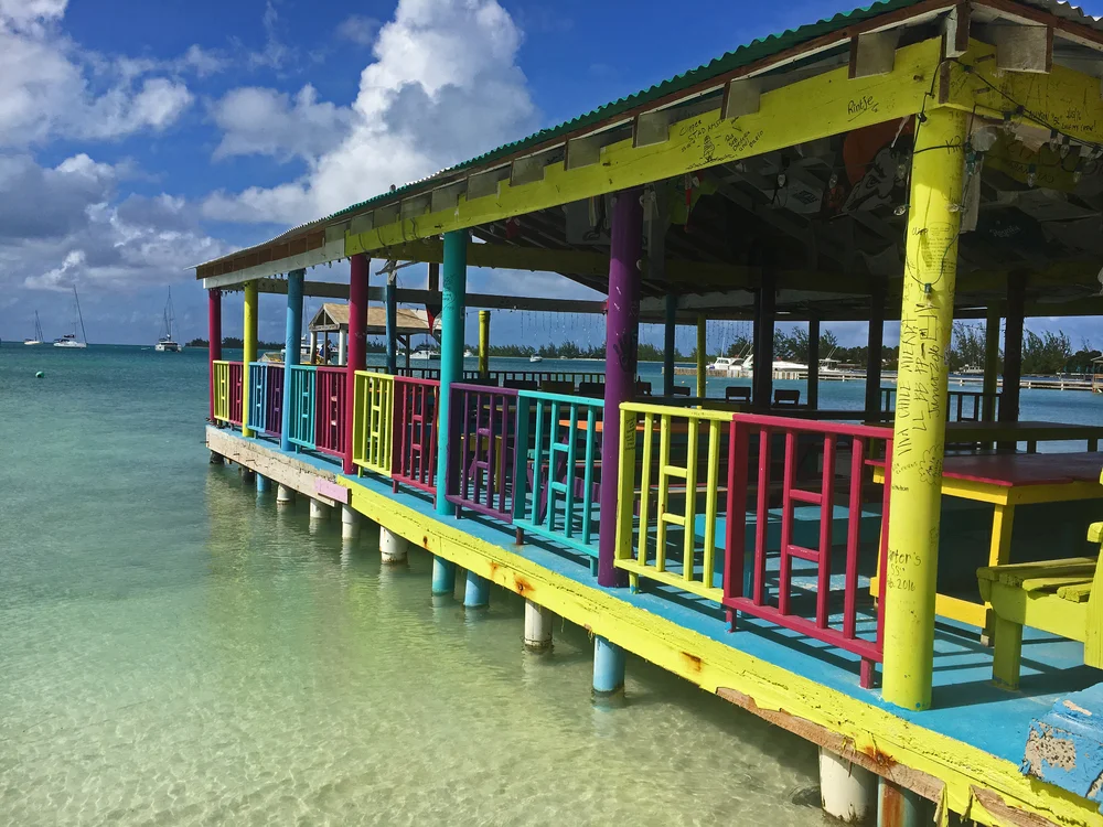 A colorful wooden pier with bright yellow, pink, purple, and blue railings extends over clear turquoise water under a partly cloudy sky. Sailboats float in the distance.