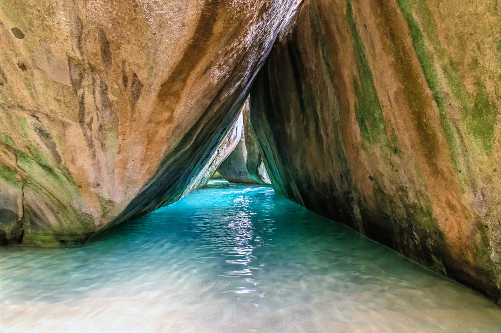Clear turquoise water flows beneath large, smooth boulders forming a narrow passageway with sunlight illuminating the opening in the distance. The rocks are streaked with shades of brown, green, and tan.