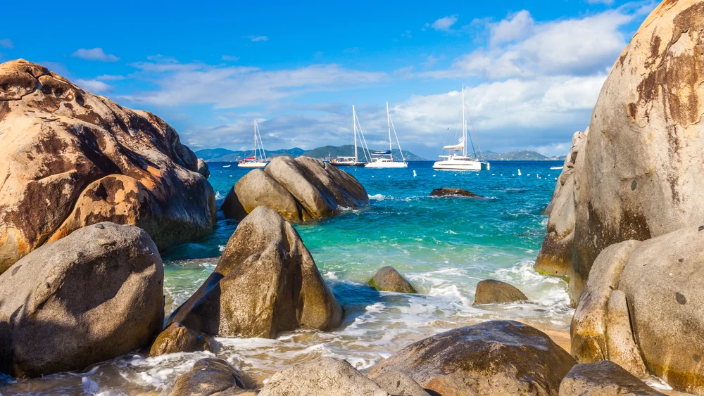Large smooth boulders frame a view of turquoise sea with gentle waves. Several sailboats float on the water under a bright blue sky with scattered clouds.