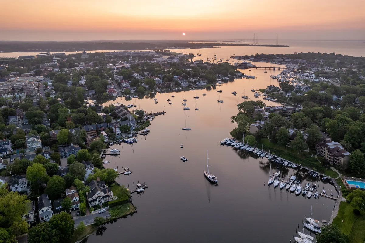 Aerial view of a coastal town at sunset, with boats docked along the calm water and houses surrounded by green trees; the sun is low on the horizon, casting a warm glow over the scene.