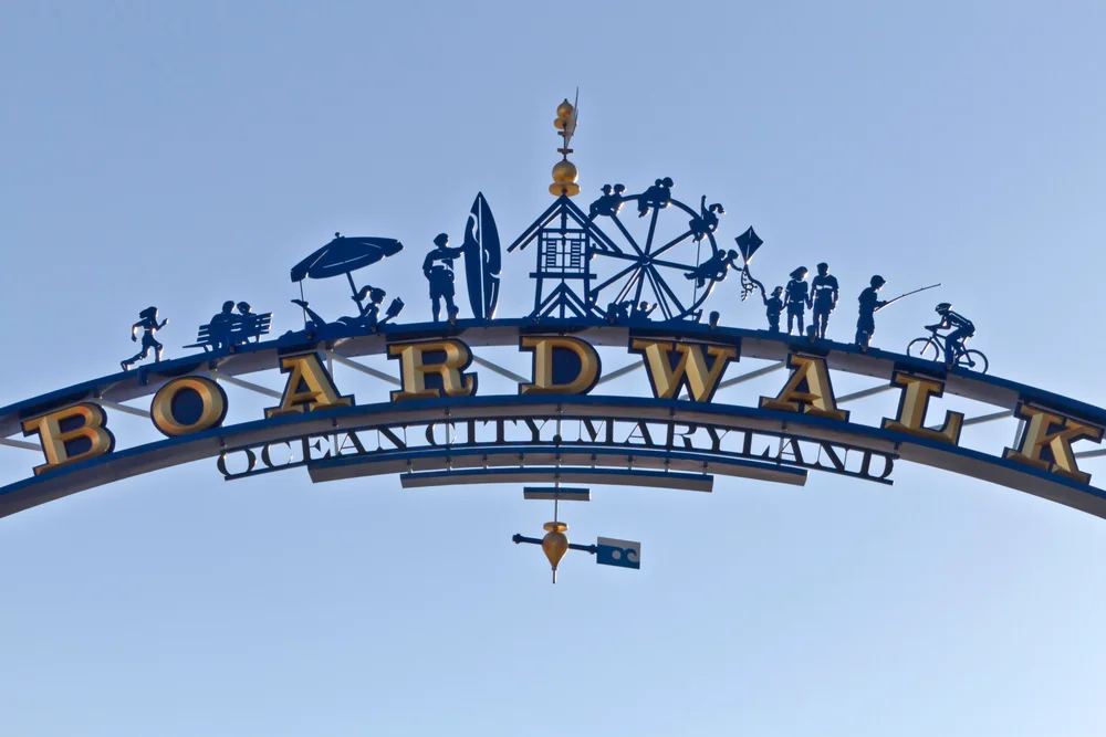 An arched metal sign reads BOARDWALK OCEAN CITY, MARYLAND with silhouettes of beach and amusement activities, including people, a surfer, Ferris wheel, and cyclist, against a clear blue sky.