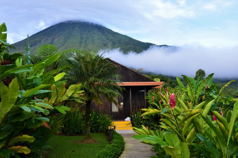 A small wooden cabin surrounded by lush tropical plants sits at the end of a path, with a misty volcano and dense greenery in the background under a cloudy sky.