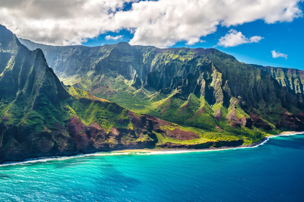 A dramatic coastline with steep, green cliffs meets turquoise ocean water under a partly cloudy blue sky, showcasing lush scenery and rugged terrain.