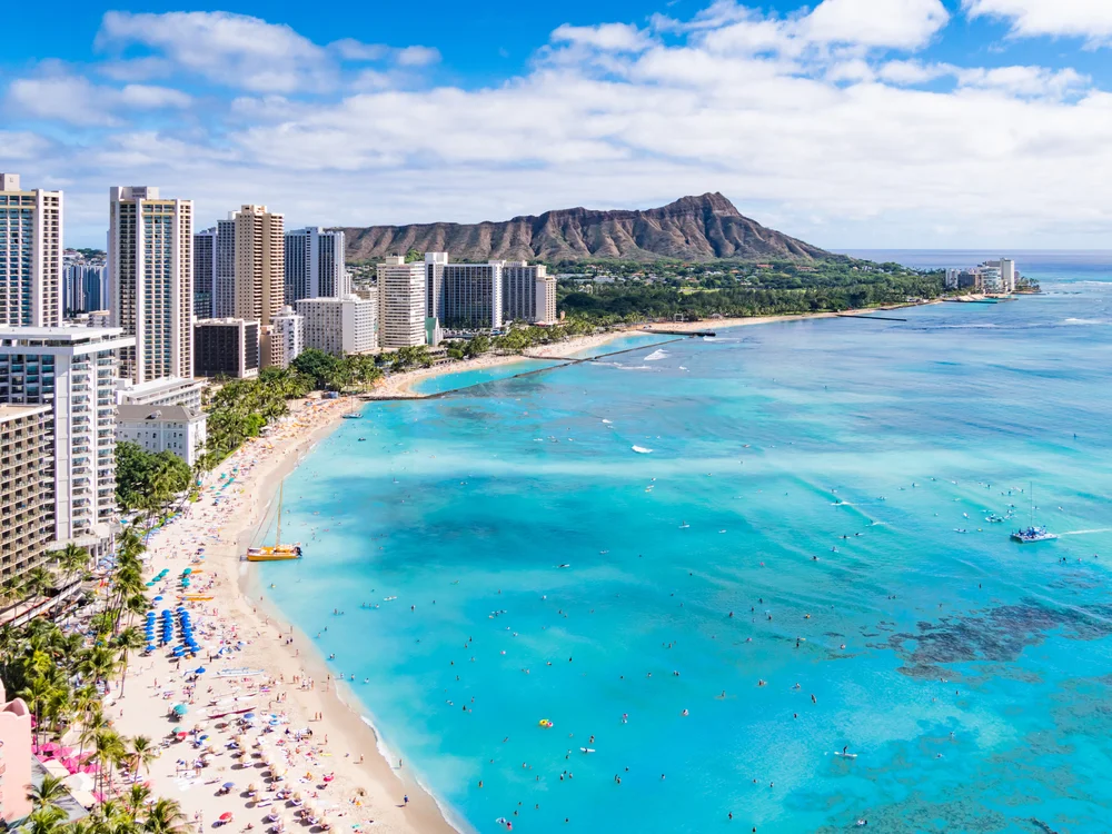 Aerial view of Waikiki Beach in Honolulu, Hawaii, with turquoise water, people swimming, sandy shore, high-rise hotels, and Diamond Head crater in the background under a partly cloudy sky.