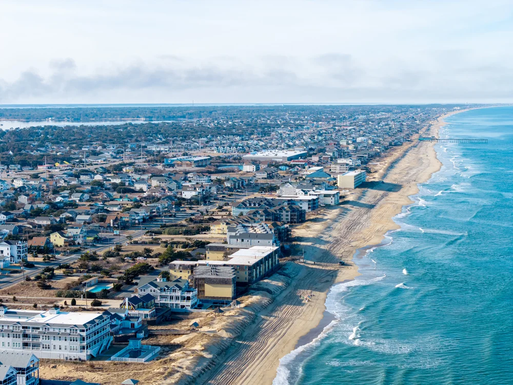 Aerial view of a coastal town with numerous buildings lined along a sandy beach, beside calm blue ocean waves, stretching into the distance under a partly cloudy sky.