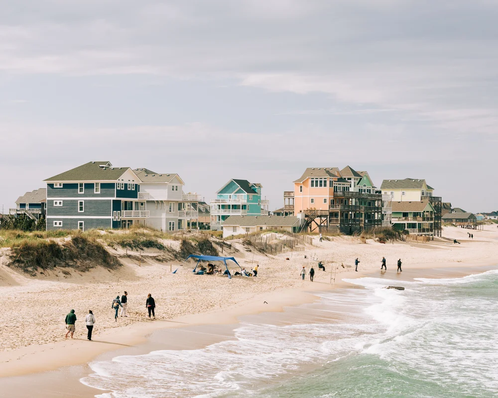 People walk along a sandy beach near ocean waves, with several colorful beachfront houses on stilts in the background under a cloudy sky. Some people are gathered near a tent on the sand.