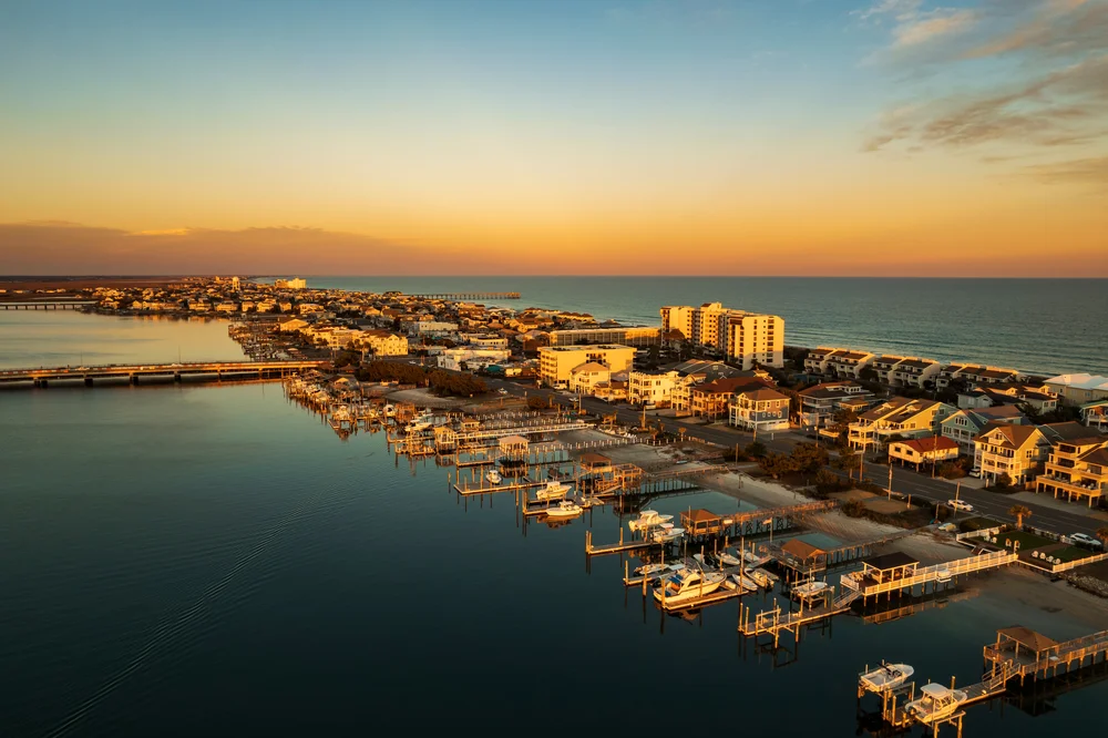 Aerial view of a coastal town at sunset, showing docks with boats, waterfront houses, and a calm ocean stretching towards the horizon under a partly cloudy sky.