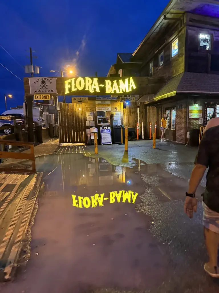 A neon Flora-Bama sign glows at night, reflected in a large puddle on the ground outside a rustic building. A few people walk nearby under the evening sky.