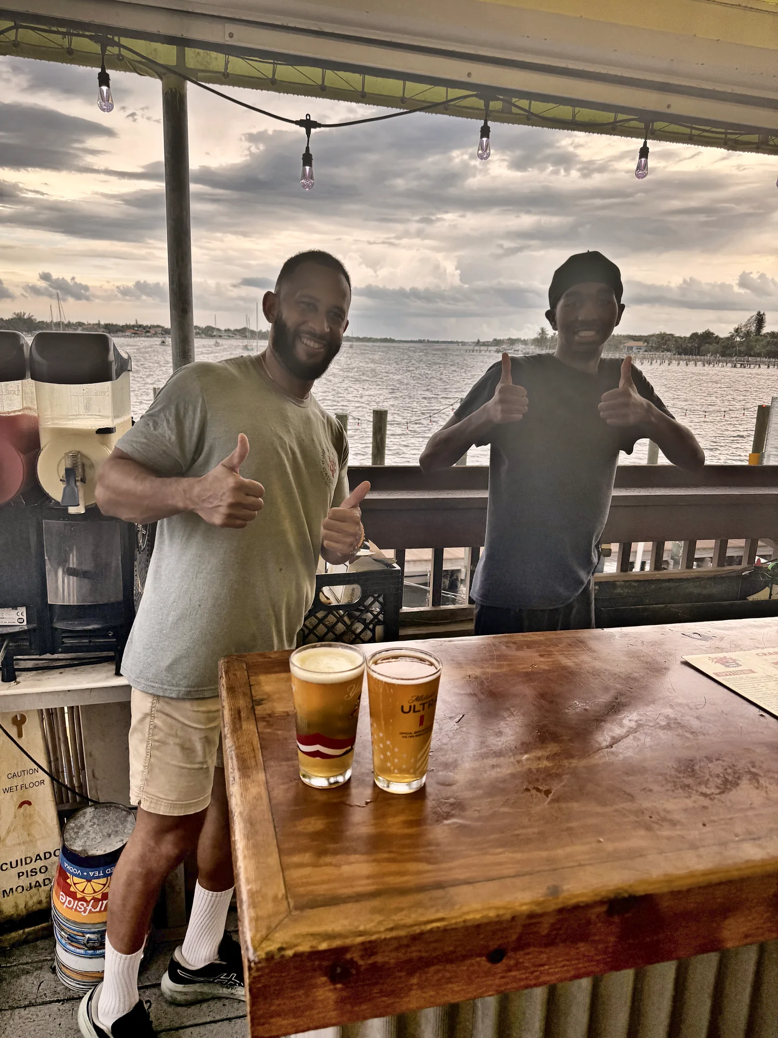 Two guests with beers and thumbs up at the wooden bar at Old Key Lime House, water behind