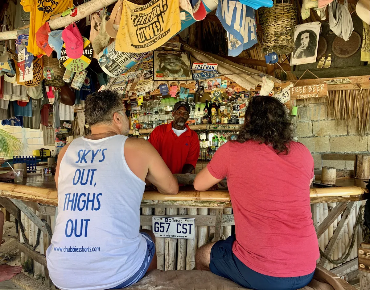 The bartender serving two guests at the bamboo bar inside Tony's Hut, T-shirts hung in the rafters
